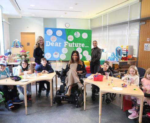 Children, teenagers and adults sitting and standing at a table of craft supplies. Two adults hold a green and blue banner that reads, 'Dear Future.'