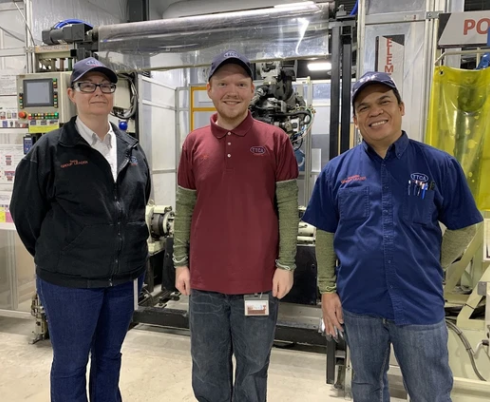 Alex stands in between two other adults in front of some factory equipment