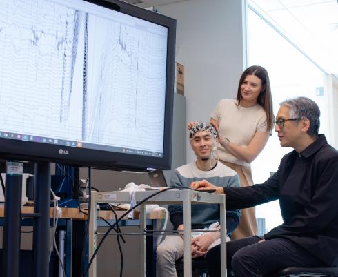 An adult sits and wears a brain computer interface headset while two other adults look at brain wave imaging.