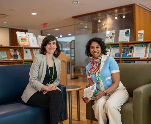Two adults sit on arm chairs in the library, surrounded by shelves of books and pamphlets.