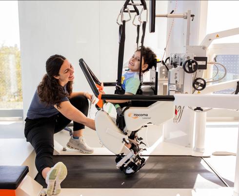 A child using a machine trying to learn how to walk, assisted by two hospital staffs