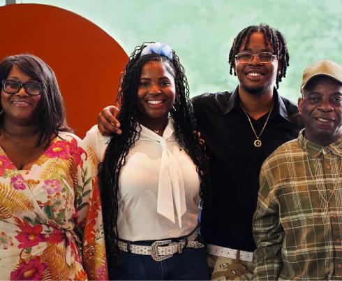 Photograph of four people standing side by side in front of a large orange circular backdrop with a green blurred background. They are wearing casual clothing, 