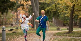 Two clowns dancing along a treed pathway holding musical instruments. They have wide smiles on their faces.