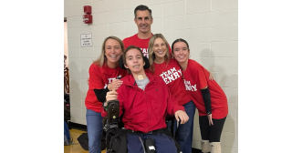 a group of family members huddled around a boy in a wheelchair device. All are wearing red shirts and dark pants. A white background behind them. Everyone is smiling at the camera.