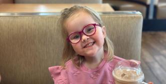 A child wearing glasses, standing in a restaurant with a cold drink in front of her